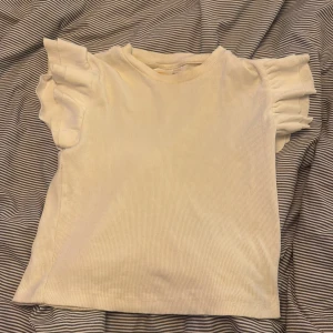 Vit volangtopp från Gina Tricot - Supersöt vit topp från Gina Tricot med ribbad struktur och volangdetaljer på ärmarna.