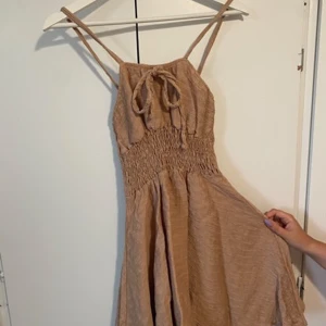 Beige sommarklänning med knyt - Supersöt beige klänning med smock i midjan och tunna axelband. Knytning framtill och lätt, luftig känsla – perfekt för varma dagar. Klänningen har en avslappnad passform och är tillverkad i ett mjukt material som känns skönt mot huden.