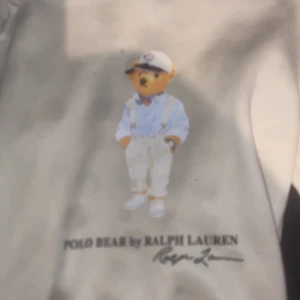 Vit hoodie Polo Bear Ralph Lauren - Vit hoodie från Ralph Lauren med ikoniska Polo Bear-trycket framtill. Klassisk huva med dragsko och lång ärm. Mjuk bomullskänsla och avslappnad passform, perfekt för en chill och stilren look.
