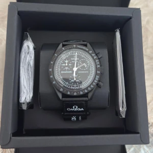 Omega x Swatch MoonSwatch Mission to the Moonphase - säljer en  klocka fick den prisent svart  Omega x Swatch MoonSwatch Mission to the Moonphase. Klockan har en rund boett, svart urtavla med vita detaljer och tachymeter-skala. Bandet är svart med Omega-logga. Tillverkad i biokeramik och levereras i originalförpackning.