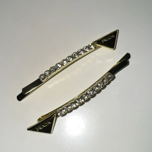 Guldiga hårspännen med strass  - Två exklusiva hårspännen  i guldmetall med glittrande strass-stenar längs hela spännet. Triangelformad detalj med P-logga i ena änden ger en lyxig och trendig vibe. Perfekt accessoar för att lyfta din look.