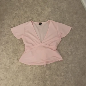 Ljusrosa omlottopp med knytband - Supersöt ljusrosa omlottopp med v-ringning och korta ärmar. Toppen har ett brett knytband i midjan som ger en snygg siluett och volangliknande nederdel. Perfekt för dig som gillar en feminin och trendig stil.