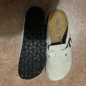 Birkenstock Boston mocka  - Birkenstock Boston i beige mocka med klassisk design och justerbart spänne i metall. Skorna har en mjuk innersula och svart räfflad yttersula för extra grepp. Perfekta för dig som gillar stilren och bekväm look.