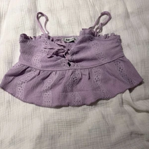 Ljuslila croppad linnetopp - Superfin ljuslila croppad linnetopp från FB Sister med smala axelband och snygg snörning framtill. Toppen har broderade detaljer och volangkant nertill som ger en söt och somrig vibe. Perfekt för varma dagar och festivaler 😍