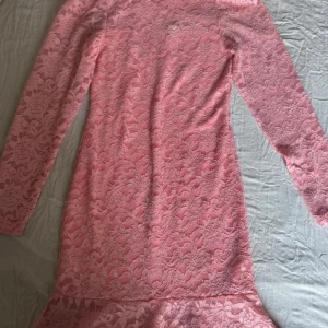 Rosa spetsklänning från Bodyflirt - Supersöt rosa spetsklänning från Bodyflirt med lång ärm och volang nertill. Klänningen har blommigt spetsmönster, knappar i nacken och figurnära passform. Perfekt för dig som vill sticka ut med en feminin och trendig look.