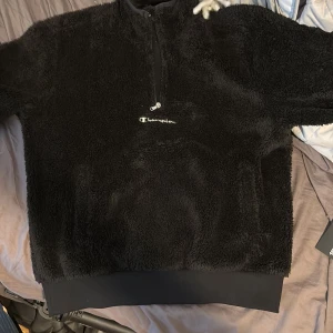Svart fleece half zip från Champion - Mysig svart fleece-tröja från Champion med broderad logga på bröstet och half zip-dragkedja. Tröjan har lång ärm och ribbad nederkant, perfekt för kalla dagar. Oversized och fluffig look som är riktigt trendig just nu.