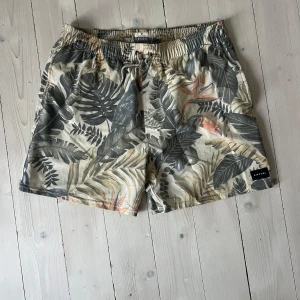 Rip Curl tropiska badshorts - Snygga badshorts från Rip Curl med tropiskt mönster i beige, grått och grönt samt detaljer i orange. Shortsen har elastisk midja med snörning och är perfekta för strandhäng eller poolen. Lätta och bekväma med coola blad och tukaner som motiv.