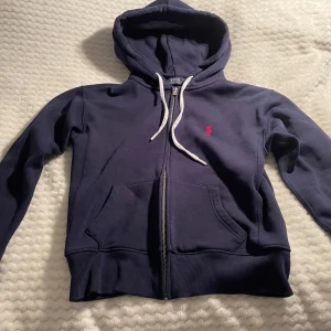 Mörkblå hoodie från Polo Ralph Lauren - mörkblå hoodie från Polo Ralph Lauren med vit snörning i huvan och dragkedja framtill. Klassisk röd logga broderad på bröstet och två fickor framtill. Pris kan diskuteras vid snabb affär 💕