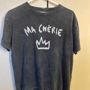 Mörkgrå t-shirt Ma Chérie Gina Tricot - Cool mörkgrå t-shirt från Gina Tricot med vit text 'Ma Chérie' och en krona på bröstet. Klassisk rund hals och korta ärmar. Materialet är mjuk bomull och passformen är normal. Perfekt för dig som gillar chill och enkel stil.