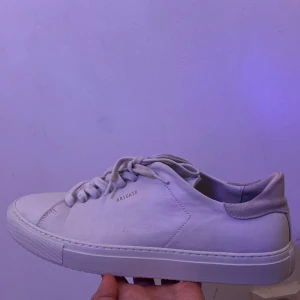 Vita sneakers från Axel Arigato - Stilrena vita sneakers från Axel Arigato med minimalistisk design. Skorna har snörning, rund tå och platt sula. Ovandelen är i slätt skinn med en detalj i beige mocka på hälen. Perfekta för dig som gillar cleana och klassiska sneakers.