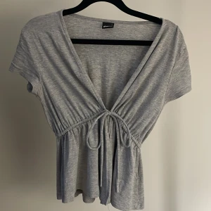 Grå omlottopp med knyt från Gina Tricot - Supersöt grå omlottopp från Gina Tricot i mjuk bomullsblandning. Toppen har korta ärmar, v-ringning och knyt framtill som ger en snygg siluett. Perfekt till jeans eller kjol! 