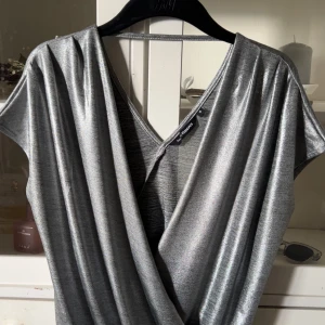 Glittrig silvertopp från KappAhl - Säljer en snygg glittrig silverfärgad topp från KappAhl. Toppen  har omlott-skärning framtill, v-ringning och korta ärmar. Perfekt för dig som vill sticka ut med en metallic look. Materialet är mjukt och följsamt syntet.