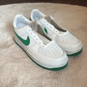 Nike Air Force 1 vit/grön sneakers - Fräscha Nike Air Force 1 sneakers i vitt skinn med gröna detaljer och grön sula. Klassisk låg modell med perforerad tå och grönt swoosh på sidorna. Perfekta för dig som gillar retrovibbar och streetstyle.