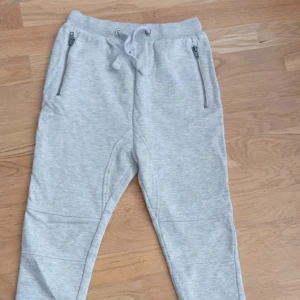 Grå mjukisbyxor med dragkedjor, 130 - Snygga ljusgrå mjukisbyxor från 157 med dragsko i midjan och dragkedjor på fickorna framtill. Byxorna har två bakfickor och är gjorda i mjukt bomullsmaterial, perfekta för chill dagar eller träning.