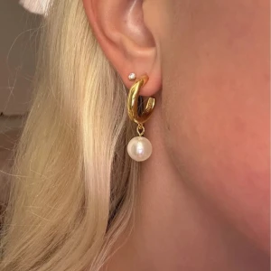 Guldiga örhängen med pärla - Säljer ett par chunky guldiga hoops med hängande rund vit pärla. Örhängena har en stilren och modern design som ger en lyxig vibe. Perfekta för dig som vill ha något som sticker ut men ändå är klassiskt. Passar till alla tillfällen och är superenkla att matcha.