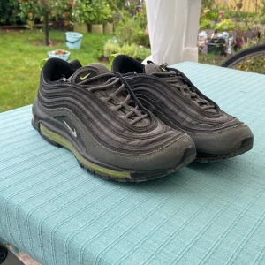 Nike Air Max 97 i mörkgrå och lime - Nike Air Max 97 sneakers i mörkgrått med limegröna detaljer och synlig Air Max-sula. Skorna har vågformade paneler, snörning och dragflik bak i neonfärg. Ovandel i syntet och textil, sula i gummi. Perfekta för dig som gillar streetwear och sportig stil.
