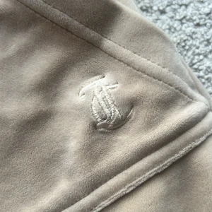  Juicy Couture byxor - Säljer ett par beige mjukisbyxor från Juicy Couture i mjuk velour. Byxorna har bred resårmidja med snörning, raka ben och fickor med lock och silverfärgade knappar bak. Ganska bra skick har några mindre fläckar. Kontakta mig om ni vill ha mer information.