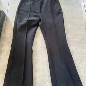 Svarta bootcut kostymbyxor Ottod'Ame - Snygga svarta kostymbyxor från Ottod'Ame med bootcut-snitt och pressveck framtill. Byxorna har hög midja, fickor i sidorna och diskreta bakfickor. Perfekta för dig som gillar stilrena och klassiska vibes. Materialet känns mjukt och har en lätt stretch.