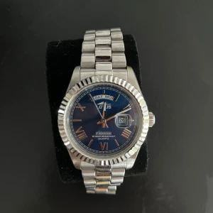 Lotus klocka - Stilren klocka från Lotus med silverfärgad länk och blå urtavla. Klockan har romerska siffror, dag- och datumvisning samt räfflad boett. Quartz-urverk och vattentålig upp till 10 bar. Perfekt accessoar för dig som gillar klassisk design med modern touch. Extra länkar följer med.