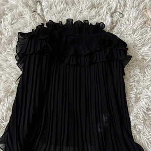 Plisserad svart volangtop från Zara - Supersnygg svart plisserad topp från Zara med volangdetaljer upptill och tunna kedjeaxelband. Luftig och flowy känsla, perfekt för att sticka ut. Toppen är ärmlös och har en elegant offshoulder-look. Ny endast testad