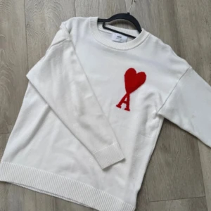 Vit stickad tröja från Ami med hjärta - Vit stickad tröja från Ami med rund halsringning och ribbade muddar. På bröstet finns ett rött hjärta och bokstaven A, vilket ger en unik och trendig look. Perfekt för dig som gillar stilrena plagg med en twist.