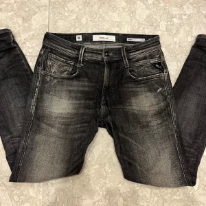 Replay Anbass svarta slim jeans - Snygga Replay Anbass jeans i svart tvätt med slitna detaljer och cool faded effekt. Slim fit-modell med klassiska fem fickor och dragkedjegylf. Perfekta för dig som gillar en modern och stilren look. Märkeslogga på fickan och bak.