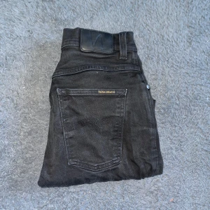 Nudie Grim Tim jeans - Säljer ett par svarta  jeans från Nudie Jeans i storlek 32/32. De har klassisk femficksdesign, svarta sömmar och Nudie-logga på bakfickan. Jeansen är tillverkade i stretchig bomull för skön passform och har dragkedja och knapp framtill. Midja 37cm. Ifall det inte finns någon bild i annonsen där jag bär plaggen jag säljer så är det för att jag ej kan ha storleken heller passformen, så kolla måtten för storlek och googla på modellnamnet för passform 😉