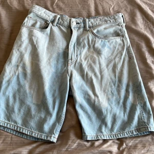 Ljusblå jeansshorts från H&M - Säljer ett par ljusblå jeansshorts från H&M i relaxed fit. Klassisk femficksmodell med bälteshällor och knappgylf. Perfekta för varma dagar och har en loose passform som ger en chill vibe. Notera det udda mönstret på dem. Det är alltså inte fläckar, utan menat att vara så. Nypris 400kr