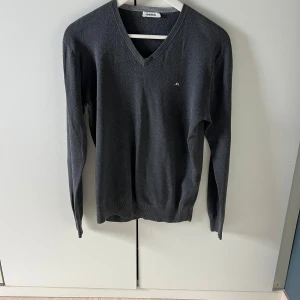 Grå v-ringad tröja J.Lindeberg  - Mörkgrå v-ringad tröja från J.Lindeberg i storlek S. Tröjan har lång ärm, liten broderad logga på bröstet och är tillverkad i ett mjukt, stickat material. Den är i okej skick och kan tänkas gå ner i pris vid snabb affär!