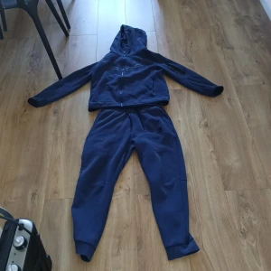 Nike tech dress - Mörkblå hoodie från Nike med dragkedja och huva. Tröjan har långa ärmar, en diskret logga på bröstet och en praktisk ficka med dragkedja på ena ärmen. Tillverkad i mjuk bomullsmix, perfekt för chill eller träning. byxorna har några skador och en fläck, se bild 3 och 4 den har också ett liiitet hål där bak. Om du vill så kan du också köpa enskilt. Om du köper enskilt så kostar byxorna 300kr enskilt och tröjan 500kr. Pris kan diskuteras!