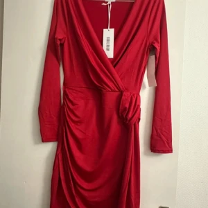 Röd omlottklänning från Bubbleroom - Snygg röd omlottklänning från Bubbleroom med lång ärm och markerad midja. Klänningen har v-ringning, draperad detalj i sidan och är tillverkad i ett mjukt, stretchigt material. Perfekt för dig som vill sticka ut med en klassisk men trendig look.