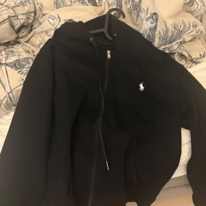 Svart hoodie från Polo Ralph Lauren - Svart hoodie från Polo Ralph Lauren med klassisk vit logga på bröstet. Tröjan har huva med snörning, dragkedja framtill och känguruficka. Perfekt för en chill och stilren look.