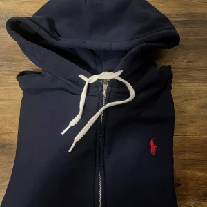 Hoodie från Polo Ralph Lauren - Snygg marinblå hoodie från Polo Ralph Lauren med dragkedja och vit snörning i huvan. Klassisk röd broderad logga på bröstet och mjukt bomullsmaterial. Perfekt för alla dagar. Passar dig som e 165-170 ca