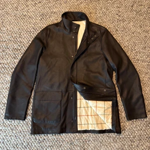 Loro piana voyager - Snygg svart skinnjacka med hög krage, stora fickor framtill och dragkedja med tryckknappar. Insidan har ett ljust rutigt foder i beige, grönt och rött. Jackan har en rak passform och klassisk look, perfekt för dig som gillar stilrena detaljer.