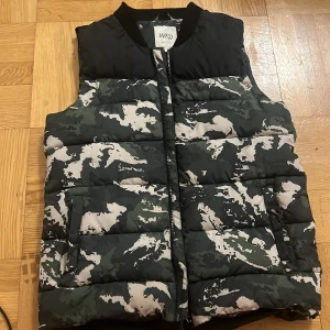 Camouflage dunväst från Warp - Säljer en fet dunväst från Warp med camouflage-mönster i svart, grått och grönt. Västen har dragkedja framtill, två fickor med knapp och hög krage. Perfekt för lager-på-lager och streetstyle.