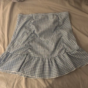 Blårutig volangkjol från Gina Tricot - Supersöt kort kjol från Gina Tricot i blått och vitt rutigt mönster. Kjolen har volang nertill som ger en extra touch och är gjord i mjukt bomullstyg. Perfekt för dig som gillar en chill och trendig stil.