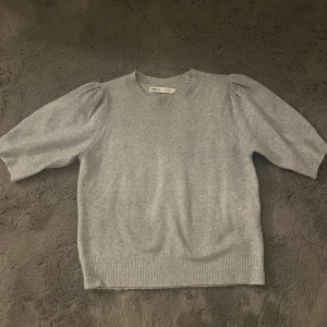 Grå stickad topp från ONLY - Supermjuk grå stickad topp från ONLY med korta puffärmar och rund halsringning. Ribbstickad kant nedtill och vid ärmslut ger en snygg finish. Perfekt för dig som gillar enkel men trendig stil.