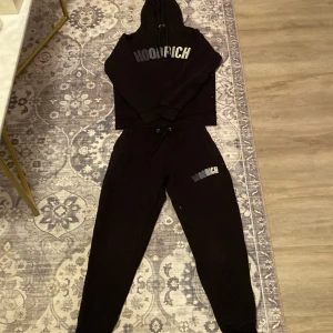 Svarta mjukisbyxor från Hoodrich - Svarta mjukisbyxor från Hoodrich med dragsko i midjan och logga tryckt på låret. Sköna och avslappnade med elastisk midja och muddar vid bensluten. Perfekta för chill eller streetwear-stil. Banden är förstörda och priset kan diskuterad. Man kan även köpa byxorna och hoodien separat Byxorna är Storlek S. Hoodien XS 