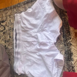 Vita sportiga shorts med randig midja - Säljer ett par vita shorts med elastisk midja och tunna svarta ränder längs linningen. De är tillverkade i mjuk bomull och har en avslappnad passform, perfekta för träning eller chill dagar.