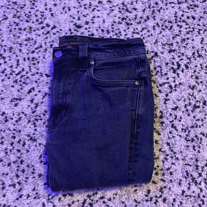 Mörkblå skinny jeans från Nudie Jeans - Säljer ett par mörkblå skinny jeans från Nudie Jeans med klassisk femficksdesign och snygga detaljer på bakfickan. Jeansen är i stretchigt denim och har en smal passform som sitter tajt längs benen. Perfekta för dig som gillar en clean och stilren look.