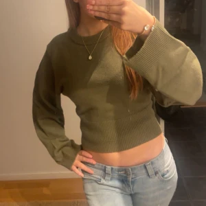 Olivgrön croppad stickad tröja - Snygg olivgrön croppad stickad tröja med breda ribbade muddar vid ärmslut och midja. Tröjan har lång ärm och en relaxed passform som ger en chill vibe. Perfekt att matcha med jeans för en avslappnad look.