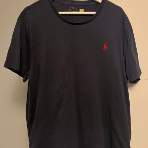 Svart t-shirt från Ralph Lauren - Svart t-shirt från Ralph Lauren med klassisk rund hals och röd broderad logga på bröstet. T-shirten är kortärmad och har en clean, stilren look som funkar till jeans eller shorts. Materialet är mjuk bomull för skön känsla hela dagen.