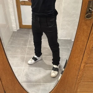 Svarta Replay Anbass jeans - Säljer ett par svarta Replay Anbass jeans med slim passform. Byxorna har klassisk femficksdesign, dragkedjegylf och diskreta detaljer på bakfickan. Perfekta för dig som gillar en stilren och modern look.