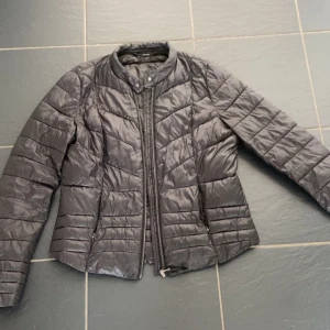 Svart tunn vår/höst jacka  - Snygg svart vår/höst jacka med quiltad design och dragkedja framtill. Jackan har en hög krage med tryckknapp och långa ärmar. Perfekt för kyliga dagar och enkel att matcha med olika outfits.