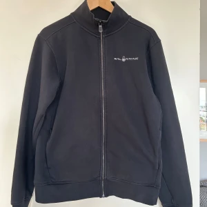 Svart ziptröja Sail Racing M - Svart ziptröja från Sail Racing i storlek M. Jackan har hel dragkedja framtill, hög krage och diskret logga på bröstet. Tillverkad i mjuk fleece som håller dig varm under kyliga dagar. Perfekt för lager-på-lager och enkel att matcha.