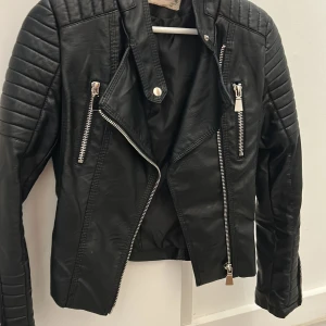 Svart skinnjacka med dragkedjor - Snygg svart skinnjacka från Wrangler med coola detaljer som silvriga dragkedjor, quiltade axlar och spännen i midjan. Jackan har långärmade ärmar och en klassisk bikerstil som ger en edgy vibe till din outfit.