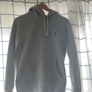 Grå hoodie från Ralph Lauren - Snygg grå hoodie från Ralph Lauren med klassisk broderad logga på bröstet. Tröjan har huva med snörning, känguruficka och långa ärmar. Perfekt för en chill och stilren look. Mjuk bomullskänsla och normal passform.