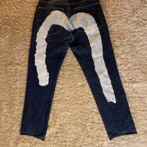 Evisu jeans med vit målad detalj, stl 30 - Säljer ett par mörkblå Evisu jeans med ikonisk handmålad vit målad båge på baksidan. Klassisk femficksmodell i kraftig denim med raka ben och snygga kontrastsömmar. Bra skick.