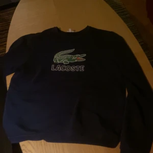 Svart sweatshirt från Lacoste - Svart sweatshirt från Lacoste med klassisk krokodil-logga och text på bröstet. Tröjan har rund halsringning och långa ärmar. Perfekt för en chill och stilren look. Passar dig som gillar sportig och enkel stil.
