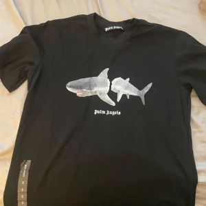 Svart t-shirt med hajmotiv från Palm Angels - Säljer en svart t-shirt från Palm Angels med tryck av två hajar på framsidan och stor vit logga på ryggen. T-shirten har normal passform och är kortärmad, perfekt för en cool och avslappnad stil.helt ny köpt för 3500